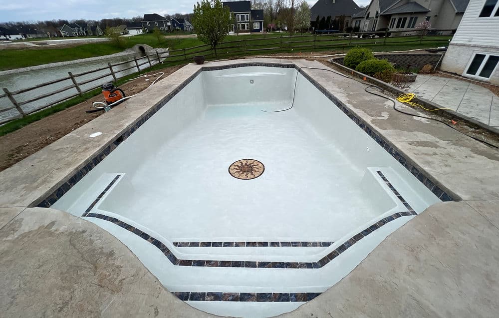 Empty in-ground swimming pool with decorative tile design, surrounded by landscaped yard.
