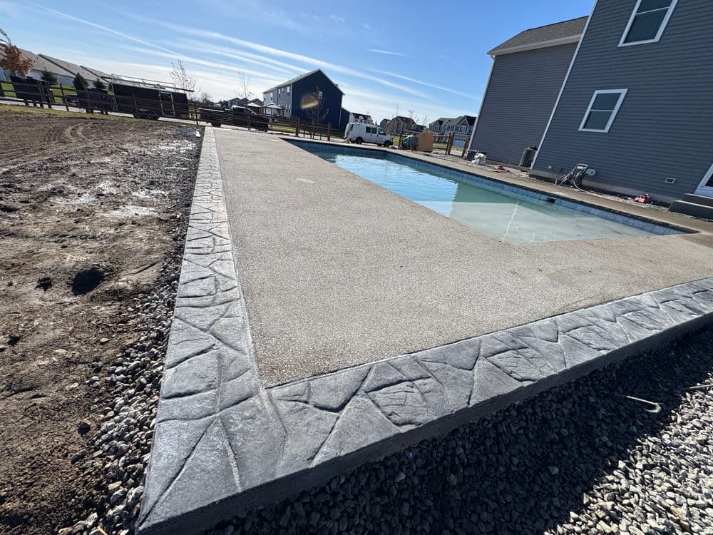 In-ground pool with textured concrete decking under blue sky and new homes nearby.