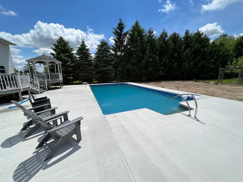 Inviting backyard pool with patio chairs, gazebo, and lush greenery under a blue sky.