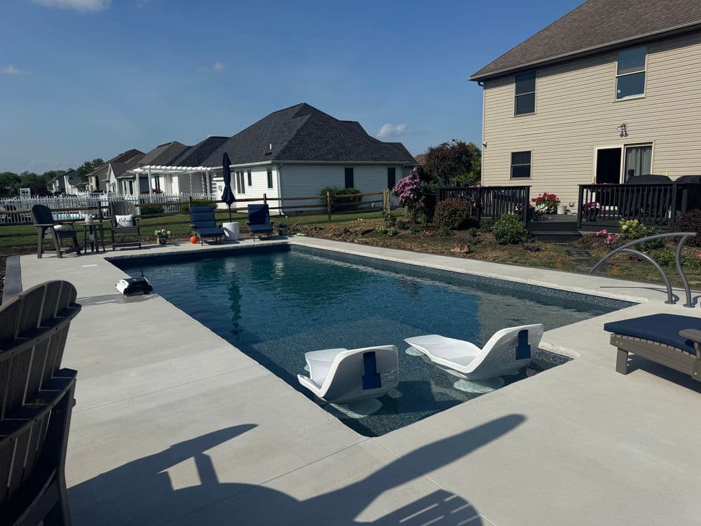 Modern backyard pool with lounge chairs, surrounded by houses and landscaping. Clear sky.