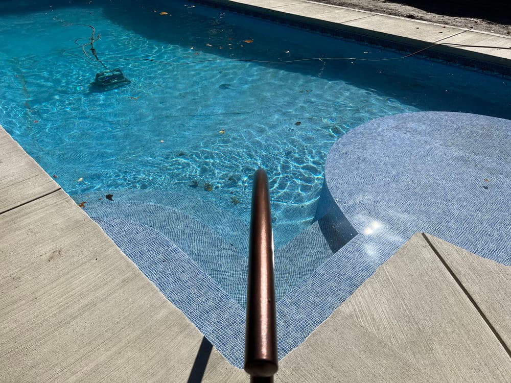 Clear swimming pool water reflecting sunlight, with a copper pool pole and textured tiles.