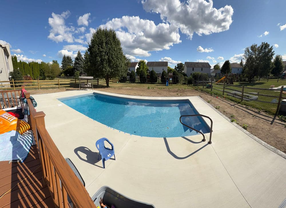 Backyard pool with clear water, surrounded by green trees and houses under a blue sky.