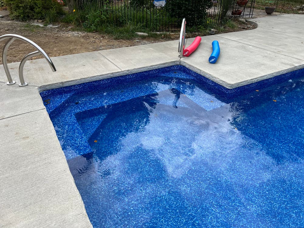 Crystal-clear swimming pool with blue tiles, pool noodles, and a landscaped yard in the background.