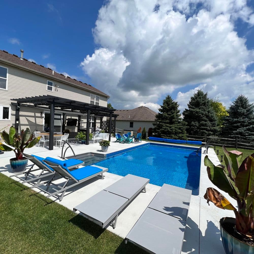 Modern backyard pool area with lounge chairs, pergola, and lush greenery under a bright sky.